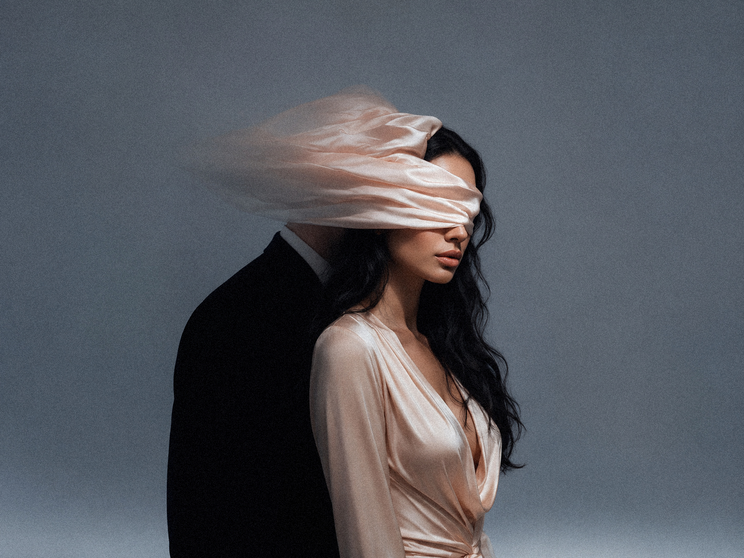 A cinematic portrait of a woman in a soft blush satin dress standing in front of a shadowed male figure, with a pale pink fabric wrapped across her eyes, symbolizing being blinded by love or emotional illusion.