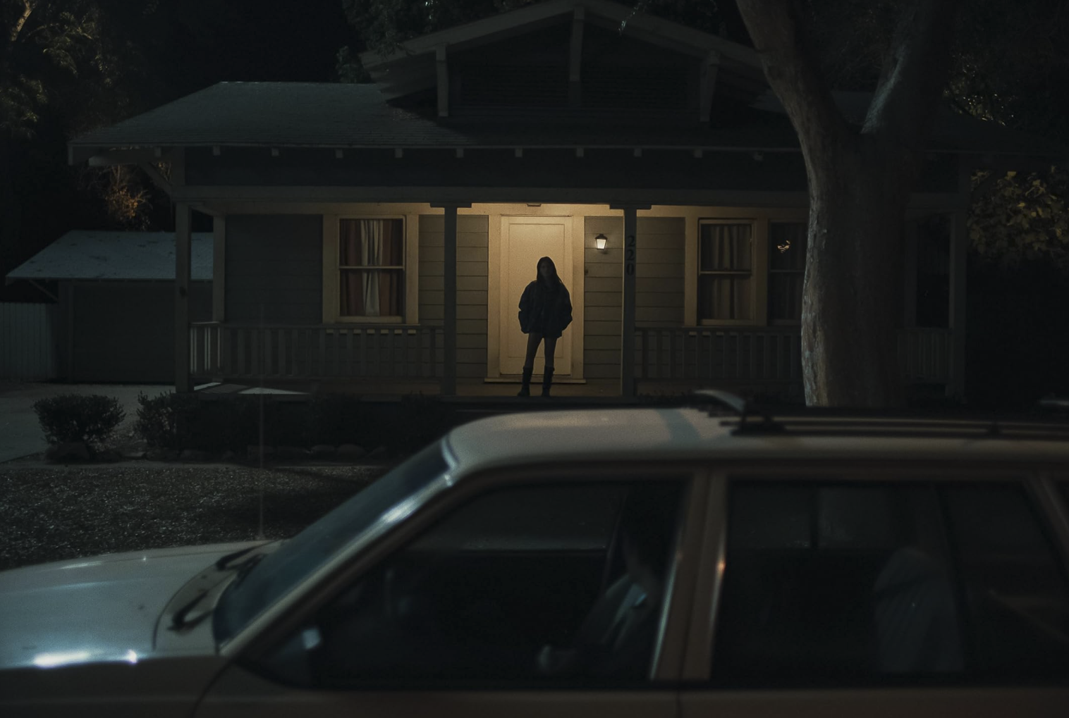 A lone young woman stands on the porch of a suburban house at night while a car is parked in the foreground, creating an eerie, suspenseful scene.