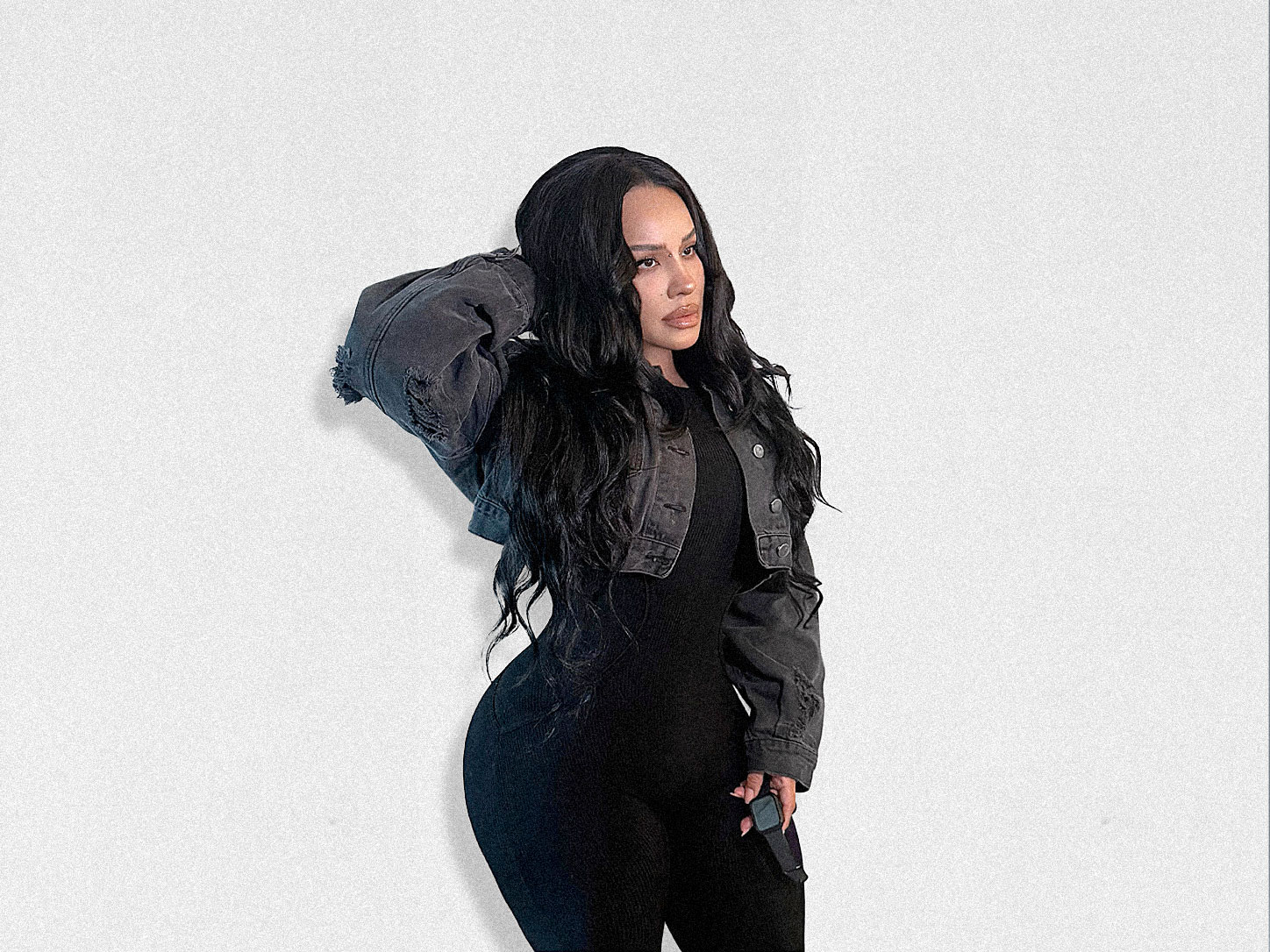 Full-body fashion portrait of a woman with long, wavy black hair wearing a fitted black outfit layered with a cropped charcoal denim jacket. She poses against a minimalist white wall with one hand in her hair, showcasing an understated monochrome look inspired by elevated everyday style.