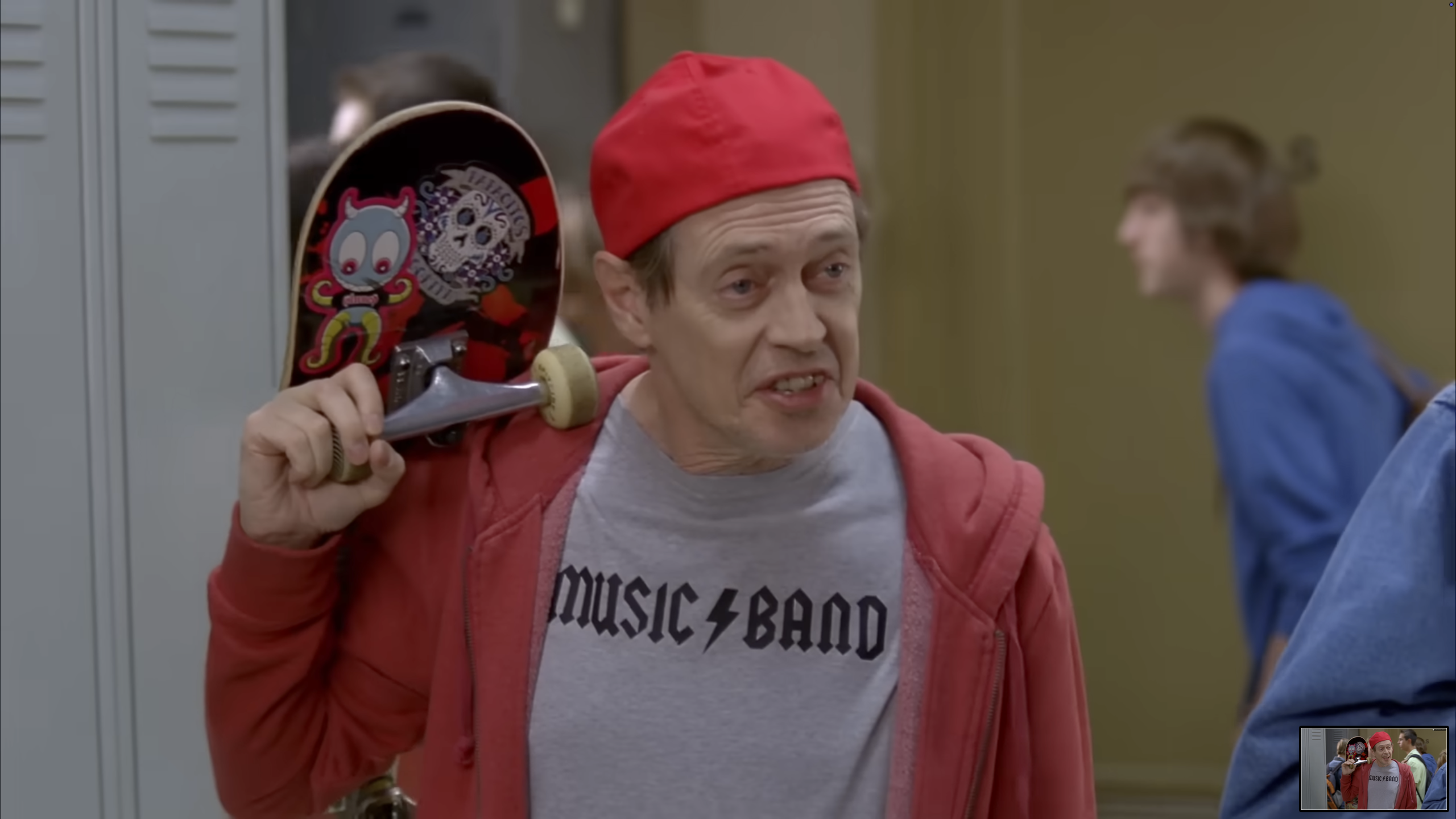 Man in a red cap and hoodie carrying a skateboard over his shoulder in a school hallway, wearing a gray “Music Band” T-shirt.